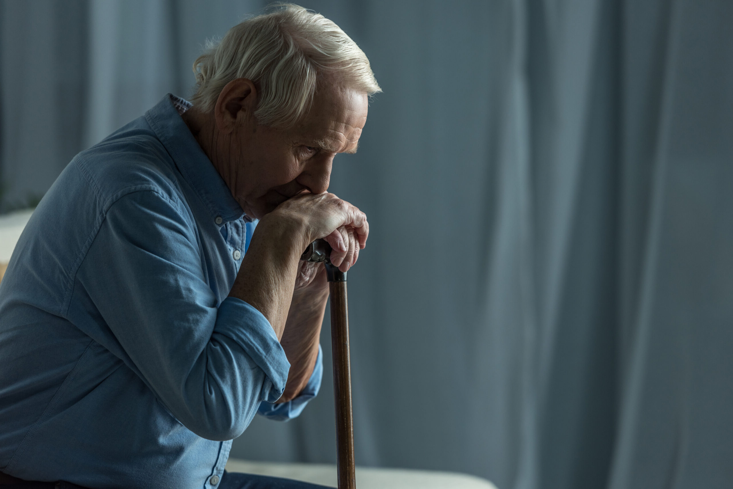 An elderly man with a sad expression sits leaning on a cane.