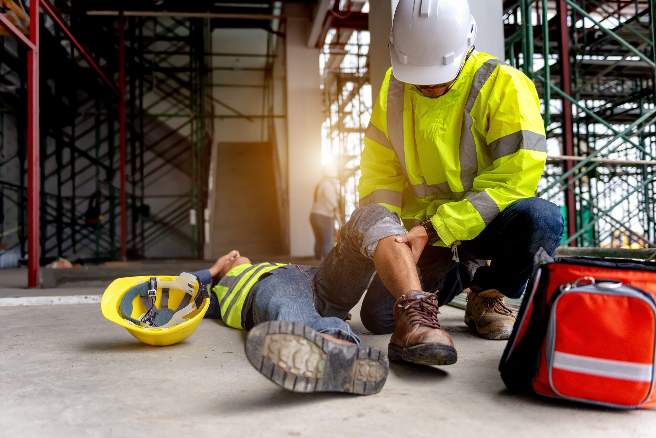 Construction worker wearing safety gear provides first aid to an injured coworker lying on the ground at a job site.