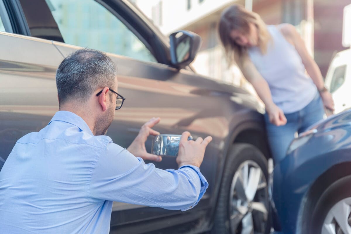 Man taking a photo of vehicle damage with his phone after a car accident, while a woman points at the damage.