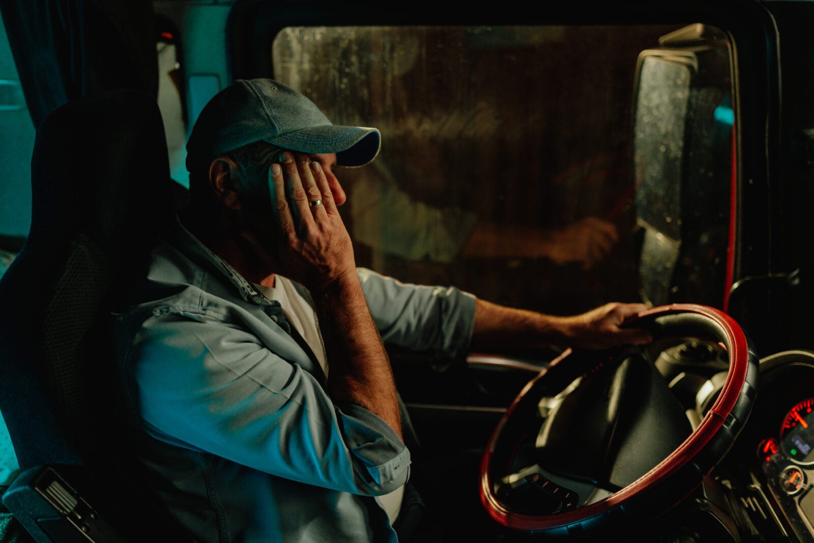 A male truck driver sits behind the wheel at night with his hand covering his face.