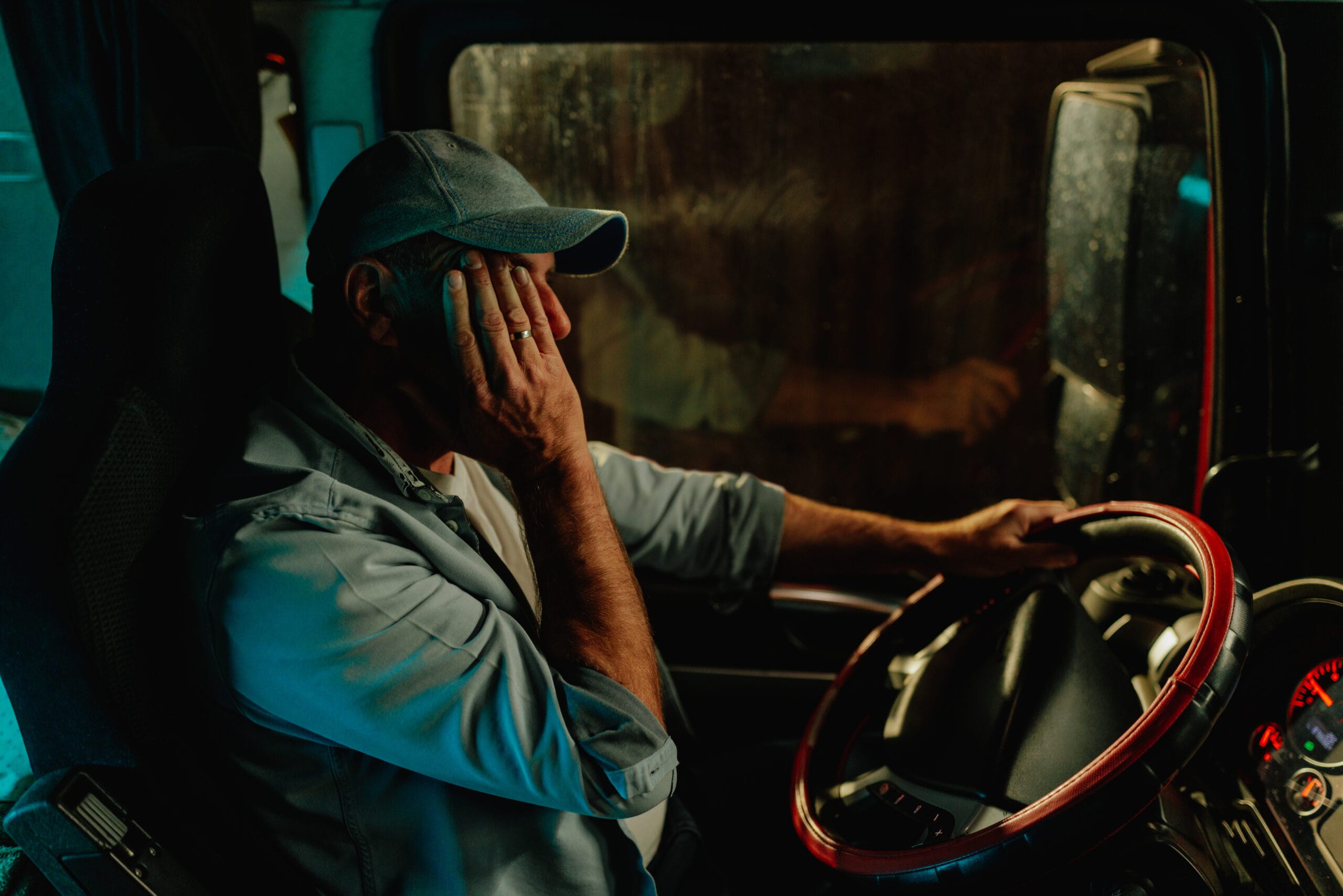 A male truck driver sits behind the wheel at night with his hand covering his face.