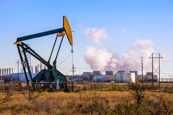 Oil pumpjack operating at an oilfield site with industrial facilities and power lines in the background.