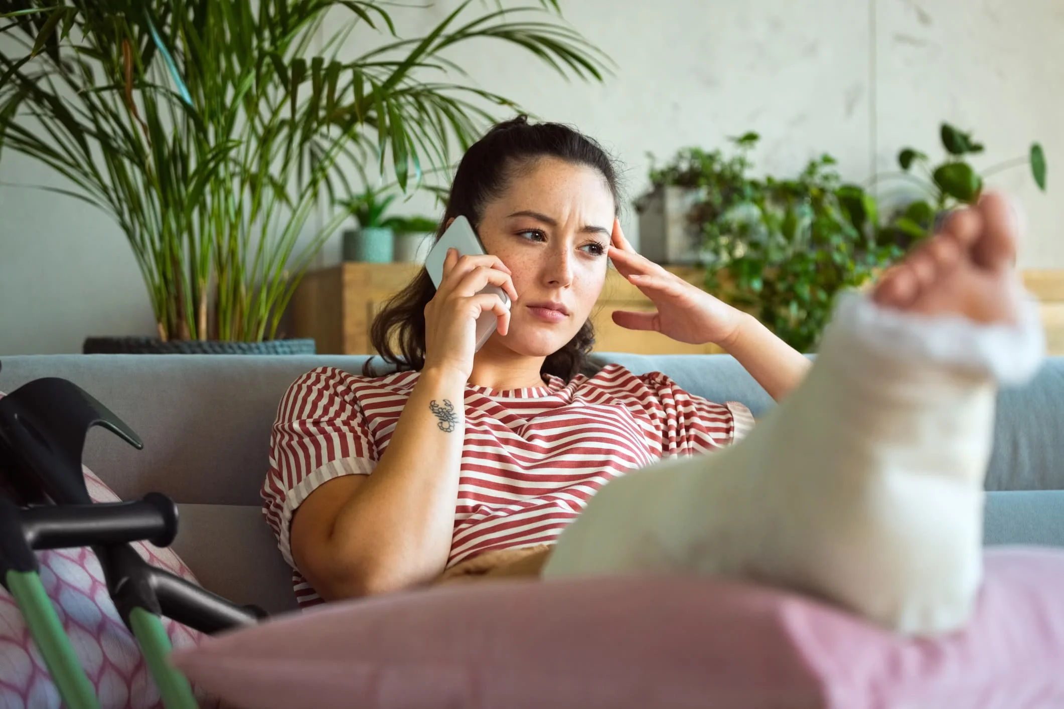 A woman with a leg cast is sitting on a couch, talking on the phone with a concerned expression.