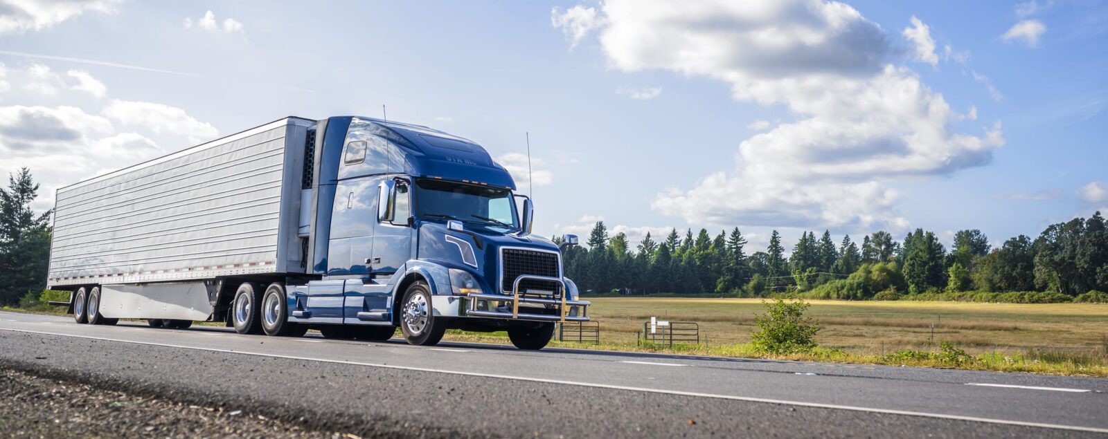 A blue semi-truck driving on a rural highway, with fields and trees lining the road.