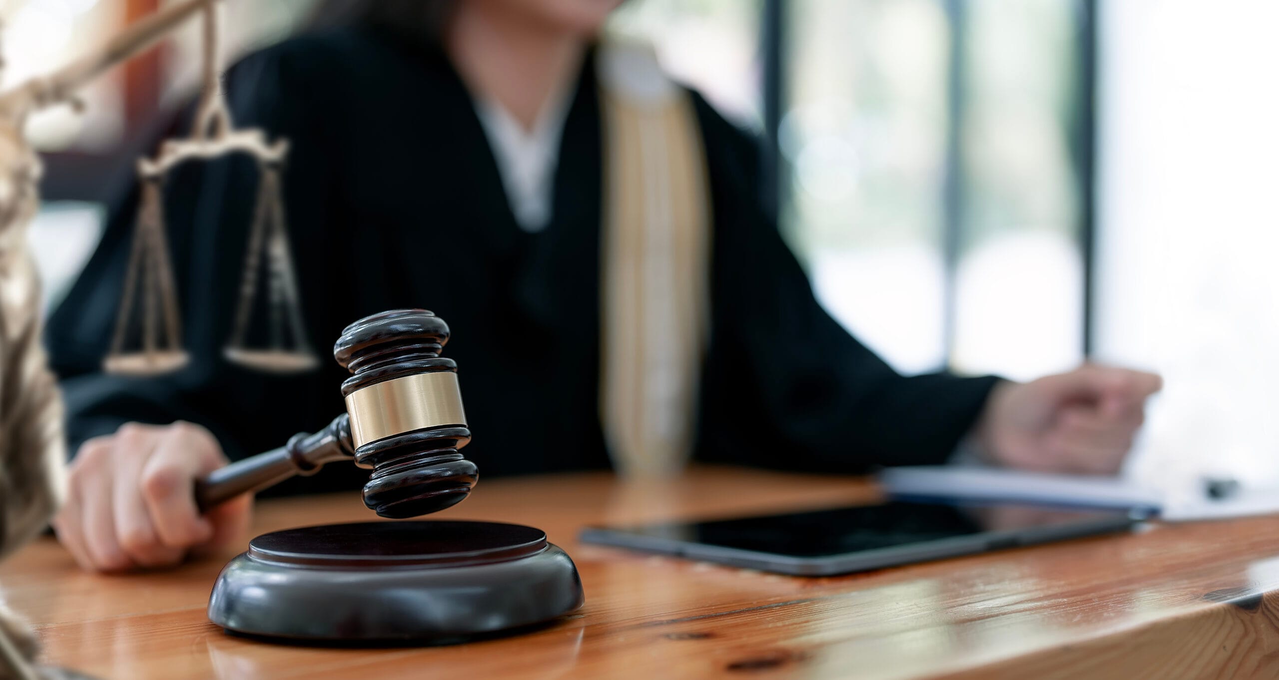 A judge in a courtroom holding a gavel near legal scales and a digital tablet.