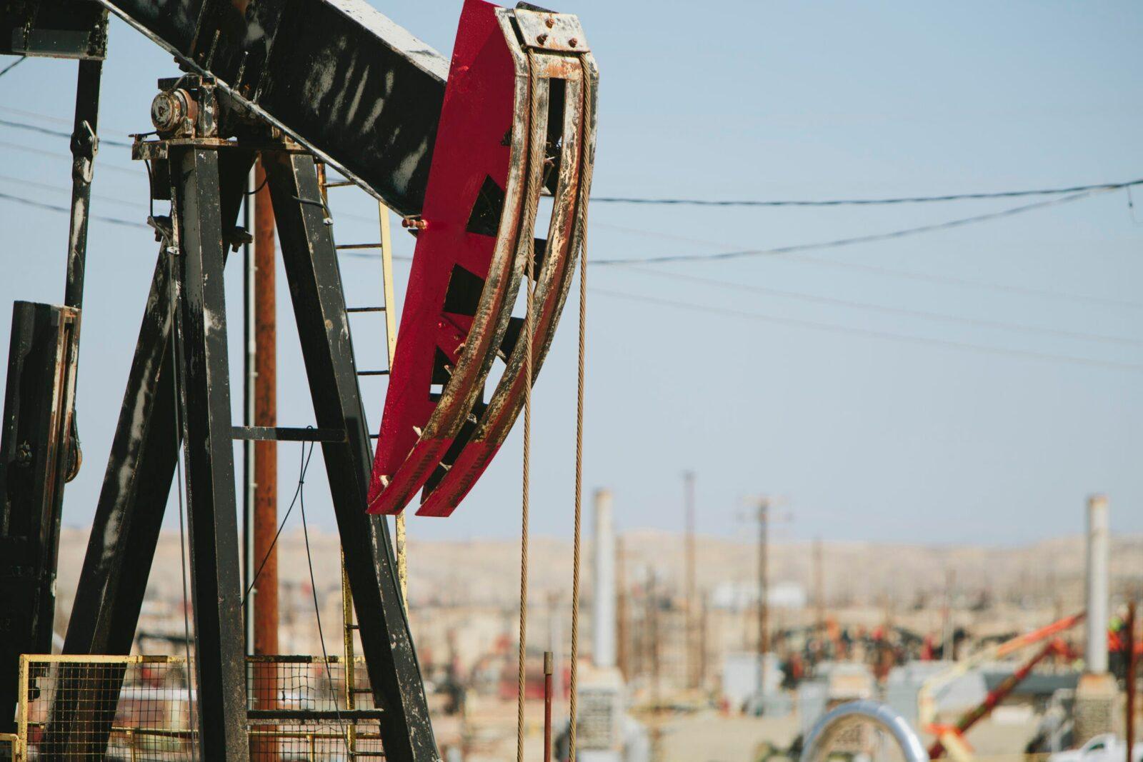 Close-up of an oil pump jack operating at an oilfield, with blurred oil rigs and industrial equipment in the background.