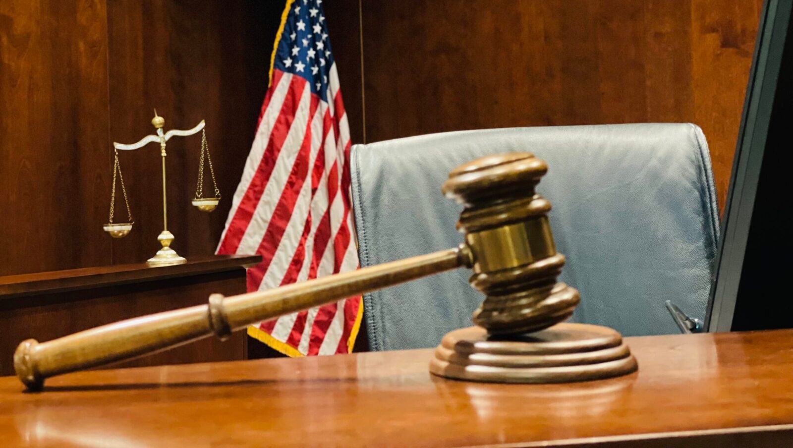 A judge's wooden gavel rests on a sound block at a judicial bench. The scales of justice and an American flag are in the background.