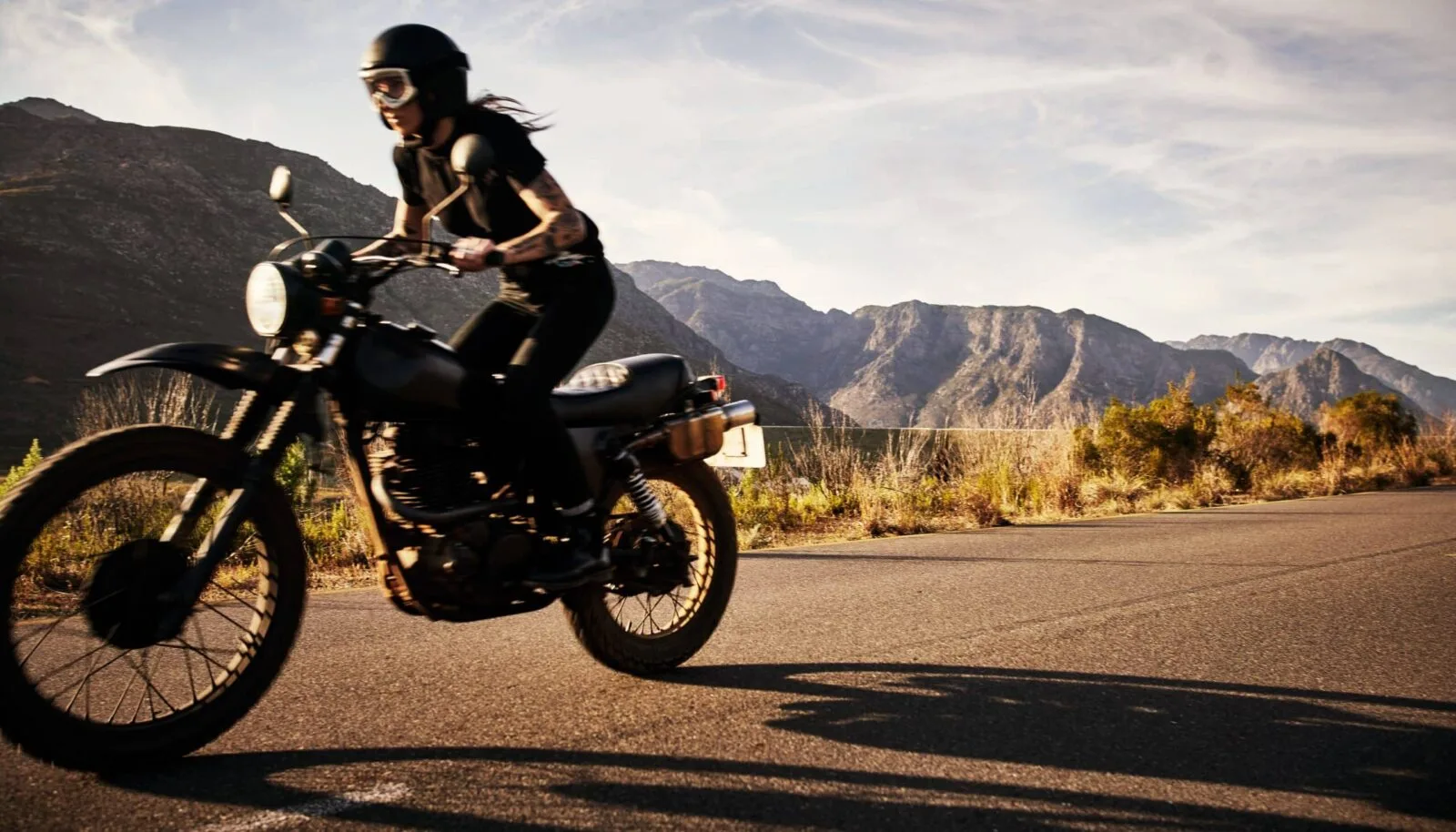 A motorcyclist wearing protective gear rides a black motorcycle on an open road with mountains in the background.