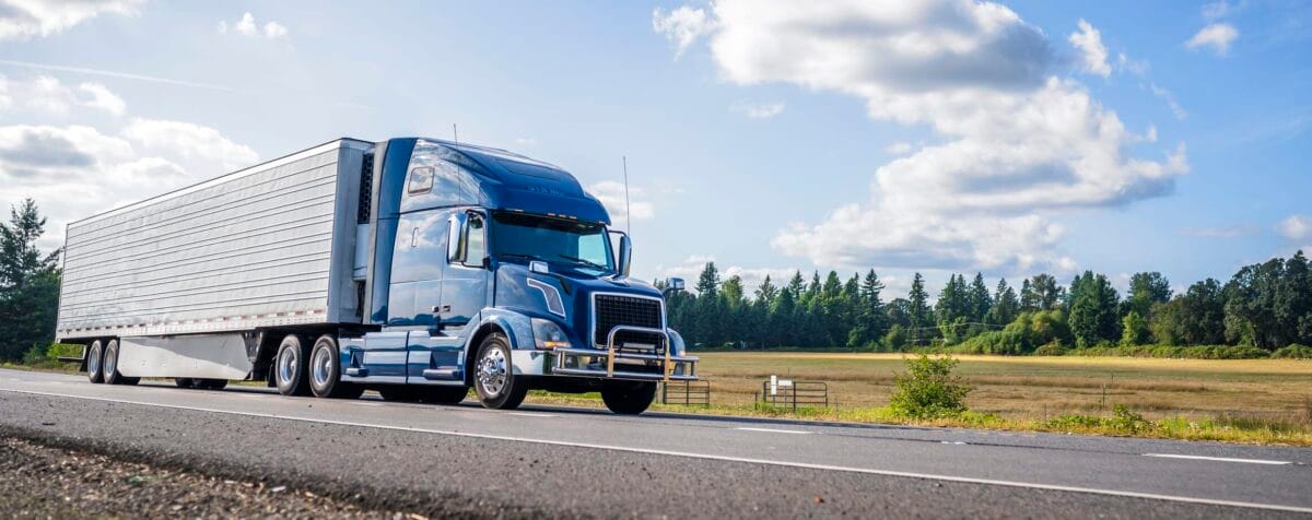 A blue semi-truck driving on a rural highway, with fields and trees lining the road.