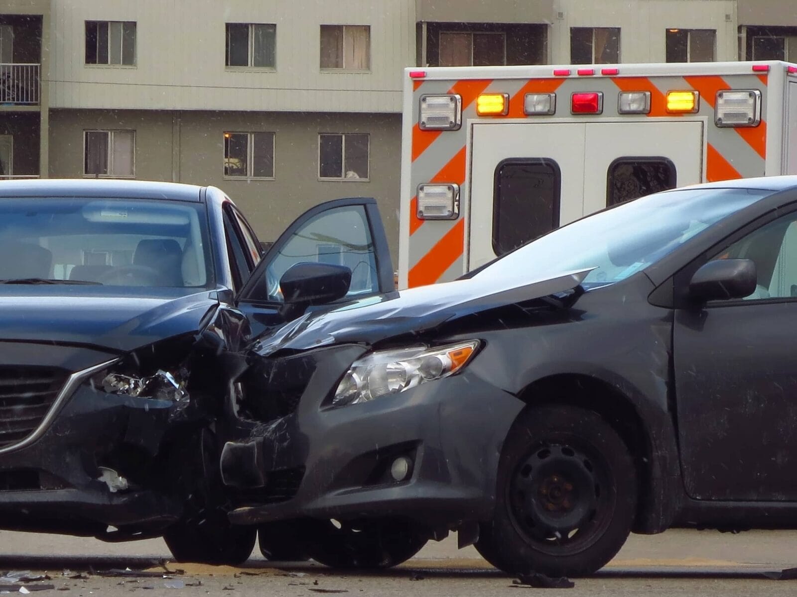 Two cars involved in a head-on collision with visible front-end damage, and an ambulance in the background.
