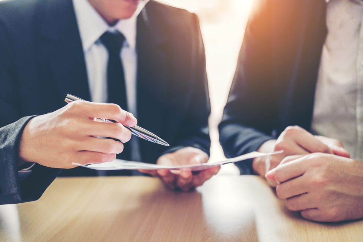 Two professionals in business attire reviewing and discussing a document at a desk, with one person holding a pen and pointing at the paper.