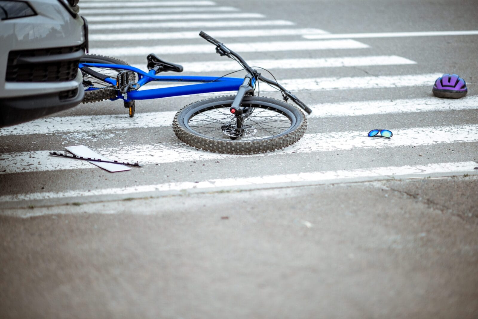 Damaged blue bicycle lying on a pedestrian crosswalk after a collision with a car, with a broken license plate, helmet, and sunglasses scattered on the road.