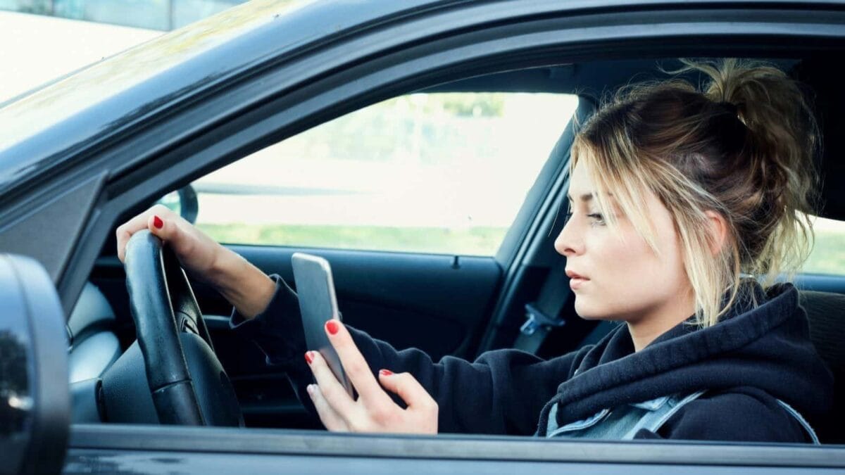 A young woman driving a car and looking at her cell phone in her hand.