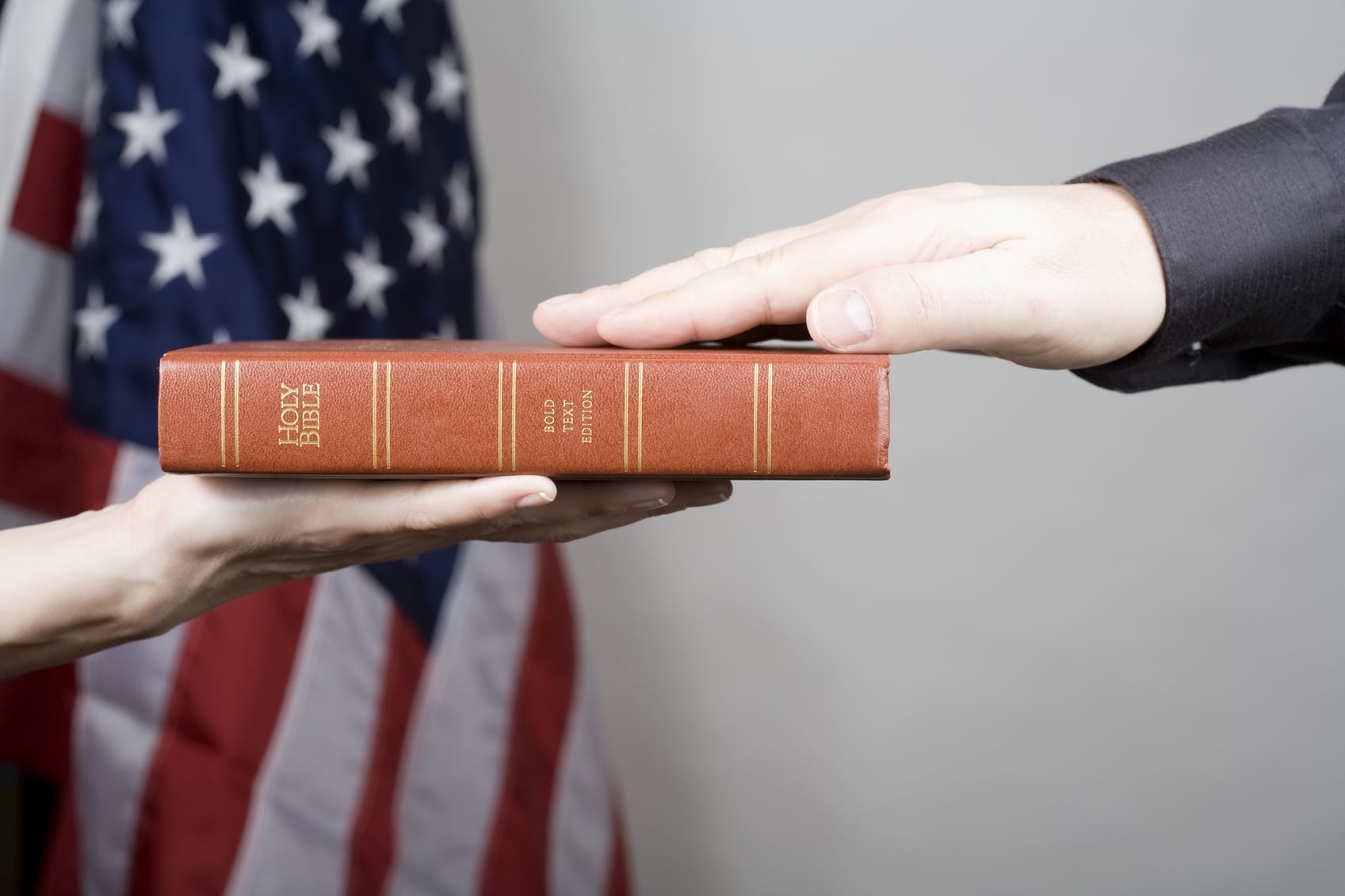 A person places their hand on a Bible held by another person, preparing to take an oath, with the American flag in the background.