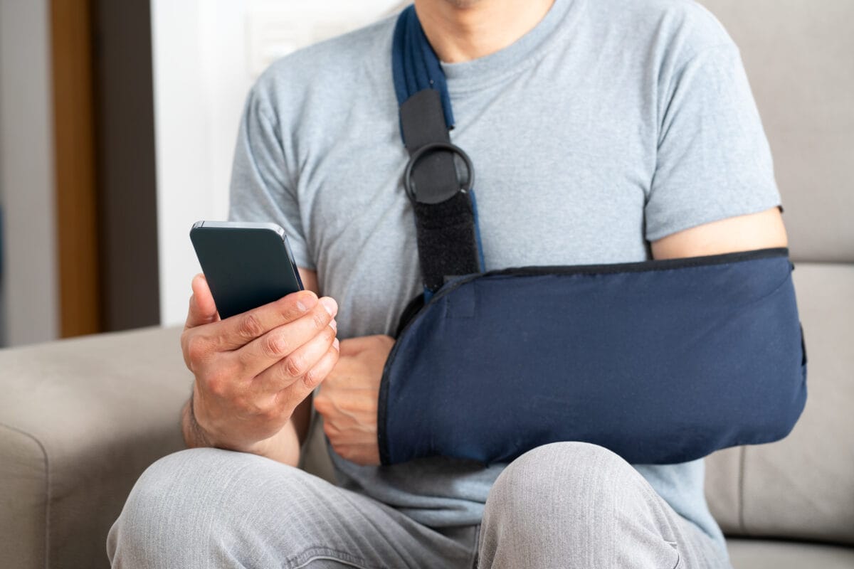 A man with his arm in a navy blue sling sits on a couch looking at his smartphone.