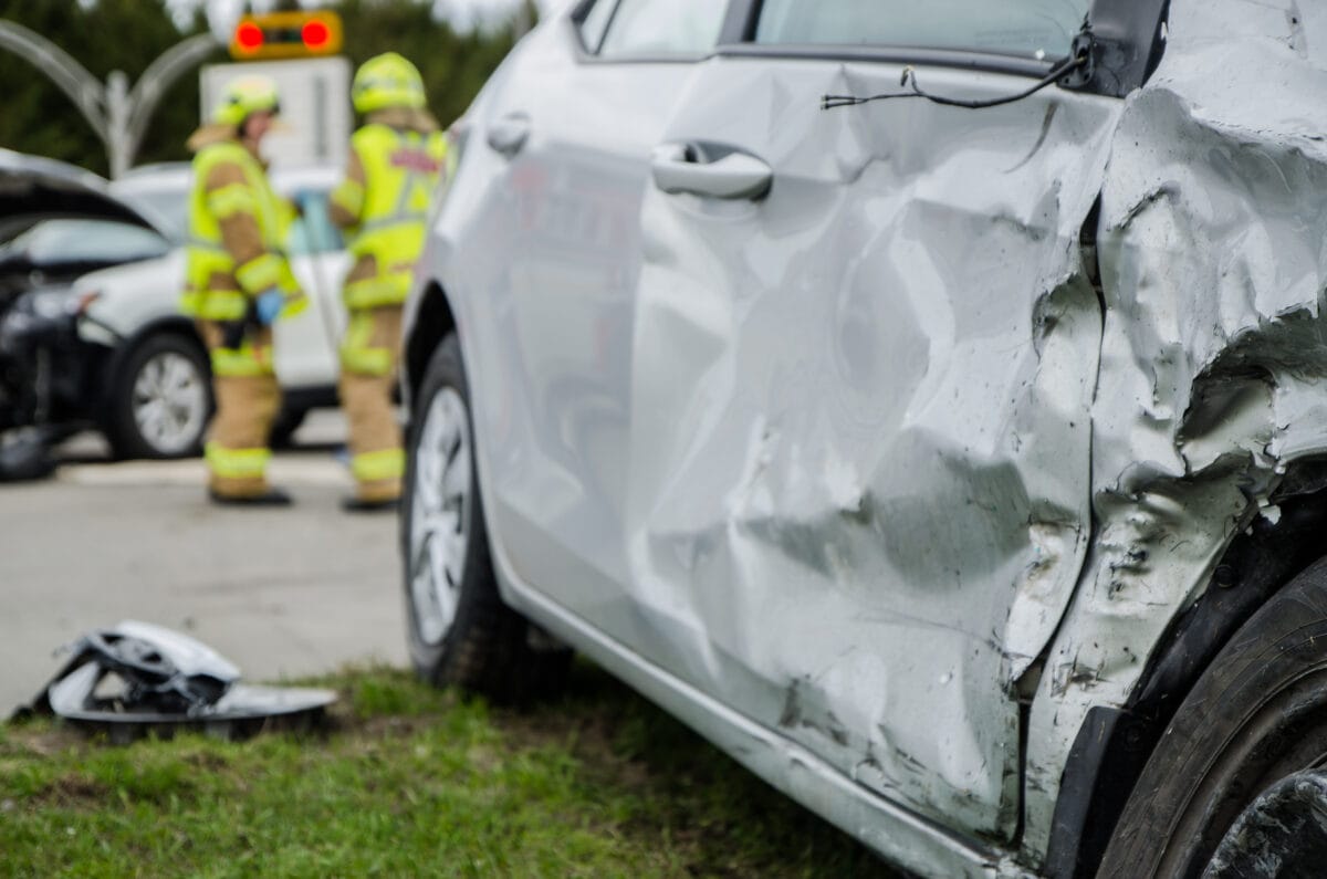 Damaged white car with severe side impact from a collision, while emergency responders stand near another crashed vehicle at an intersection.