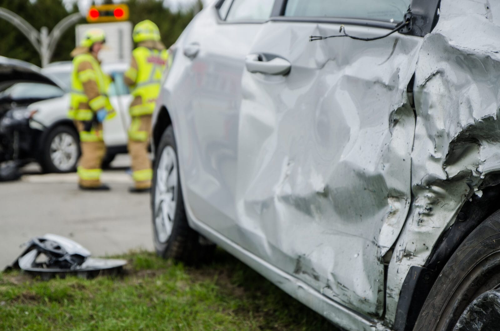 Damaged white car with severe side impact from a collision, while emergency responders stand near another crashed vehicle at an intersection.