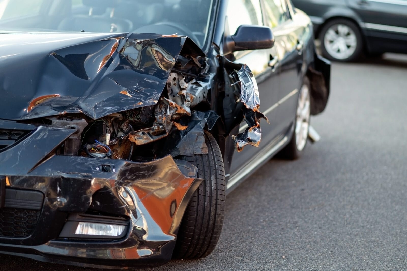 Close-up of a black car with severe front-end damage after a collision, showing crumpled metal and exposed components on a roadway.